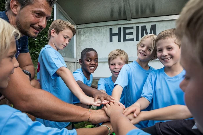 Image of a group of kids stacking hands in the centre of them all with an adult to their left, demonstrating teamwork